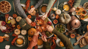 Group of people having Thanksgiving day dinner. Top view of table set with traditional Thanksgiving food and pumpkin decoration and people’s hands clinking glasses.